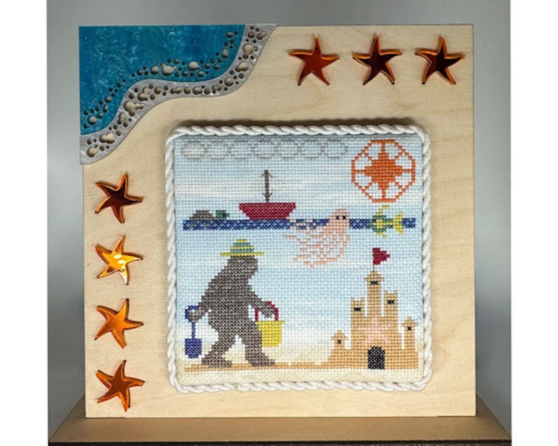 May include: A cross-stitch embroidery framed in a light wood frame with a white rope border. The embroidery depicts a brown bear wearing a blue hat and carrying a bucket, a sandcastle, a seagull, and a red boat. The frame is decorated with orange star-shaped embellishments.