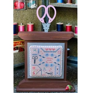 May include: A wooden sewing display with a cross-stitch design that reads "stitching mends the soul." The display features a pair of pink scissors and is surrounded by spools of thread in various colors. A strawberry is at the base.