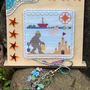 May include: A cross-stitch embroidery of a bear on the beach with a bucket and shovel, a sandcastle, and a boat. The embroidery is framed with a white rope trim and is displayed on a wooden stand with a blue and white wave design. The stand has four orange star accents. A pair of rainbow-colored scissors and a charm bracelet with sea-themed charms are also pictured.