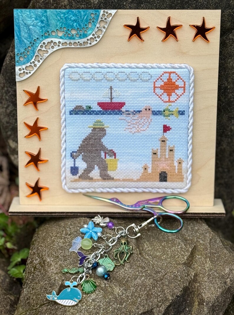 May include: A cross-stitch embroidery of a bear on the beach with a bucket and shovel, a sandcastle, and a boat. The embroidery is framed with a white rope trim and is displayed on a wooden stand with a blue and white wave design. The stand has four orange star accents. A pair of rainbow-colored scissors and a charm bracelet with sea-themed charms are also pictured.