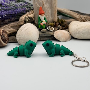 May include: Two green lizard keychains with black eyes are displayed. A small gnome figurine with a red hat and green coat is in the background, along with seashells and driftwood. The keychains are connected to a silver keyring.