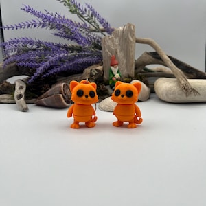 May include: A pair of orange cat-shaped earrings. Each earring features a small, standing cat with black eyes and a round head. The earrings are set against a backdrop of driftwood, lavender, and seashells.