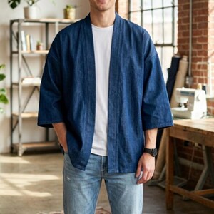 May include: A dark blue denim kimono jacket with three-quarter sleeves. The jacket is worn over a white t-shirt and paired with light blue jeans. The model is standing in a workshop setting with a brick wall and wooden shelves.