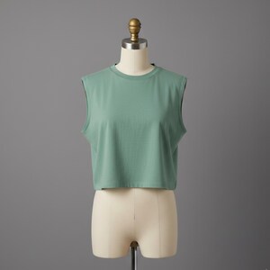 May include: A sleeveless, cropped top in a muted green hue. The top has a crew neck and a simple, minimalist design. It is displayed on a mannequin against a gray backdrop, showcasing its casual style and fit.