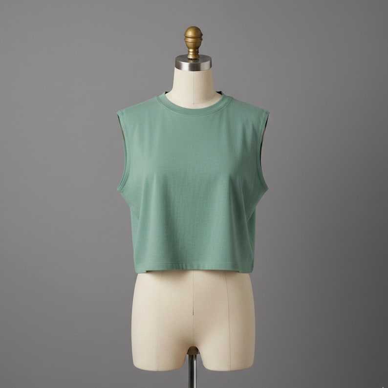 May include: A sleeveless, cropped top in a muted green hue. The top has a crew neck and a simple, minimalist design. It is displayed on a mannequin against a gray backdrop, showcasing its casual style and fit.
