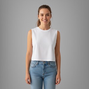 May include: A white sleeveless crop top with a crew neck. The top is made of a soft, opaque fabric. The model is wearing light blue jeans. The background is a solid gray.