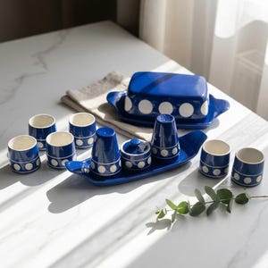 Può includere: Un set di utensili da cucina in ceramica blu con pois bianchi, che include un burriera rettangolare con coperchio, sei tazze piccole, un set sale e pepe e un piccolo contenitore con coperchio, il tutto disposto su un lungo vassoio blu curvo.