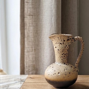 Op de afbeelding: Een bespikkelde keramische kan met een afgeronde basis en een handvat. De kan heeft een lichtbeige kleur met donkerbruine vlekken en een donkerdere bruine basis. Hij staat op een houten oppervlak.