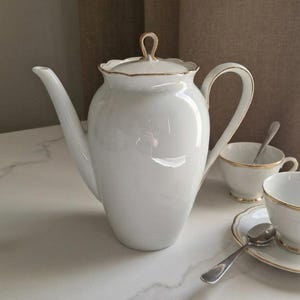 Puede incluir: Juego de té de porcelana blanca con ribete dorado. El juego incluye una tetera, dos tazas de té, platillos y cucharas. La tetera tiene un pico y asa curvos, y una tapa con asa dorada. Las tazas y platillos tienen ribete dorado.