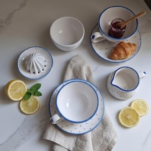 May include: A white ceramic breakfast set with blue polka dots and trim, including a teacup, saucer, bowl, creamer, and a citrus juicer. A croissant, jam, and lemon slices are also present, arranged on a marble surface.