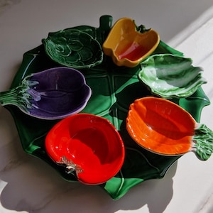 Op de afbeelding: Een grote groene, bladvormige serveerschaal van keramiek met vijf kleinere, groentevormige kommetjes op het oppervlak. De kommetjes hebben de vorm van een artisjok, een gele paprika, een paarse aubergine, een rode tomaat en een oranje wortel.