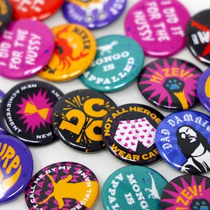 May include: A collection of colorful round buttons with various designs and text. The buttons feature a variety of colors, including pink, black, blue, and yellow. Some buttons have text like "I DID IT FOR THE NUSSY" and "NOT ALL HEROES WEAR CAPES".