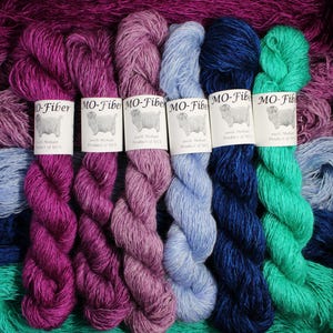 May include: A collection of colorful mohair yarn skeins in shades of purple, pink, blue, and green. Each skein has a label that reads "MO-Fiber 100% Mohair Product of USA". The yarn is arranged in a visually appealing display.
