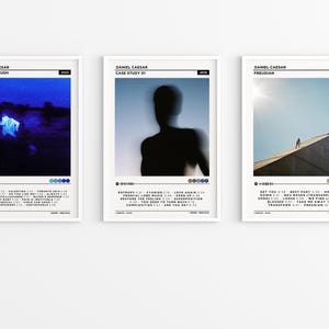 May include: Three framed art prints featuring album art for Daniel Caesar. The prints have white frames and include album titles, track listings, and the year of release. The first print has a blue-toned image, the second a silhouette, and the third a person on a building.