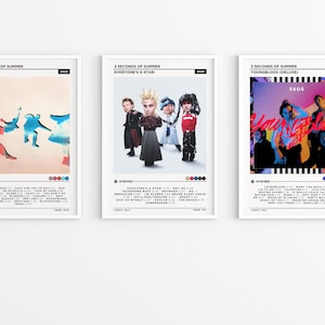 Puede incluir: Tres carteles enmarcados con portadas de álbumes de la banda 5 Seconds of Summer. Los carteles muestran portadas de álbumes con texto y gráficos coloridos. Los carteles se exhiben en una pared blanca.