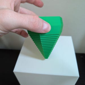 May include: A bright green, geometric object with a ridged, conical design. The object is held above a white, square-shaped box. The object's shape is reminiscent of a stylized tree or a decorative element.