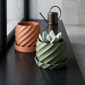 Op de afbeelding: Twee cilindrische plantenpotten met een spiraalvormige ribbeltextuur en bijpassende schotels, één in terracotta oranje en één in saliegroen, met een lichtgroene vetplant, vergezeld door een kleine donkere metalen gieter.