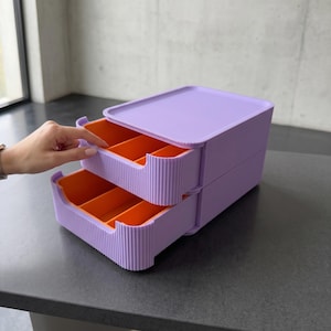 Könnte beinhalten: Ein zweistufiger Schreibtisch-Organizer mit hellvioletter Außenseite in gerippter Textur und leuchtend orangefarbenen Innenfächern. Das Gerät verfügt über einen flachen Deckel und zwei ausziehbare Schubladen mit je drei Fächern.