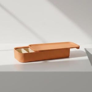 Könnte beinhalten: Eine rechteckige Aufbewahrungsbox in Terrakotta-Farbe mit passendem Deckel. Die Box hat eine gerippte Textur und ist teilweise geöffnet, wodurch ein beigefarbenes Innere sichtbar wird. Der Deckel ist leicht geöffnet und liegt auf der Seite der Box. Die Box steht auf einer weißen Oberfläche.