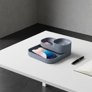 Peut inclure: Un organisateur de bureau bleu ardoise avec une texture extérieure côtelée, doté d'une base plate pour un smartphone et de deux compartiments surélevés pour de petits objets. Il contient un smartphone, un stylo et des trombones, avec un stylo et un carnet.