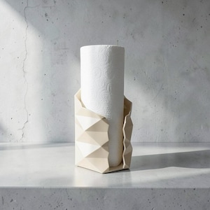 Könnte beinhalten: Ein cremefarbener Küchenrollenhalter mit einem geometrischen, facettierten Außendesign. Der Halter verfügt über eine vertikale, kantige Struktur, die eine Standardrolle weißer Küchenpapiertücher mit geprägtem Muster umschließt.