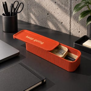 Könnte beinhalten: Ein rechteckiges, orangefarbenes, geripptes Kunststoff-Etui mit einem Schiebedeckel, der den Schriftzug "keep going" in goldener Schrift trägt. Das Etui ist teilweise geöffnet und gibt den Blick auf ein beiges Innenfach mit silbernen Büroklammern und einem schwarzen Stift frei.