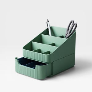 Op de afbeelding: Een groene bureau-organizer met meerdere compartimenten en een uitschuifbare lade. Een witte pen en een grijze schaar zijn in de organizer geplaatst. De organizer heeft een geribbelde textuur en is ontworpen voor kantoorbenodigdheden.