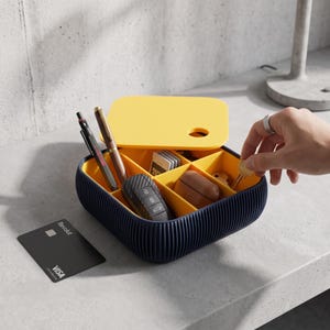 Op de afbeelding: Een vierkant, donkerblauw opbergbakje met geribbelde zijkanten en een geel interieur verdeeld in vier vakken. Het bakje bevat twee pennen, een autosleutel, een klein bruin doosje en geheugenkaarten. Een geel deksel ligt er half open bovenop.