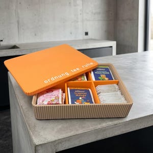 Könnte beinhalten: Eine rechteckige, beige Tee-Aufbewahrungsbox mit einem orangefarbenen Deckel, der den weißen Schriftzug ordnung. tee. ruhe. trägt. Die Box hat vier Innenfächer mit verschiedenen Teebeuteln, darunter Meßmer Tropical Mango und Minze-Honig.