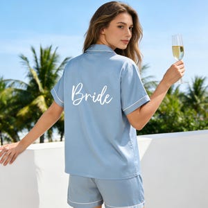 Puede incluir: Conjunto de pijama azul claro con ribete blanco. La parte superior de manga corta tiene la palabra "Bride" en blanco en la espalda. El conjunto incluye pantalones cortos a juego. Una persona sostiene una copa de champán.