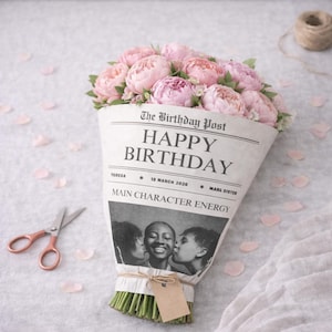 Puede incluir: Un ramo de peonías rosas envuelto en papel de periódico con la inscripción "HAPPY BIRTHDAY", la fecha "12 MARZO 2026" y "MAIN CHARACTER ENERGY". En el fondo, unas tijeras de color oro rosa y un ovillo de hilo.