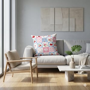 May include: A square cushion with a patchwork design featuring cherries, roses, and hearts in pink, blue, and white squares. The cushion is placed on a white sofa in a living room setting. The cushion adds a touch of charm to the room's decor.
