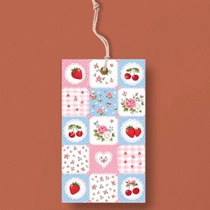 May include: A rectangular tag with a patchwork design featuring strawberries, cherries, and roses on a blue and pink background. The tag is made of paper and has a natural twine string attached at the top.