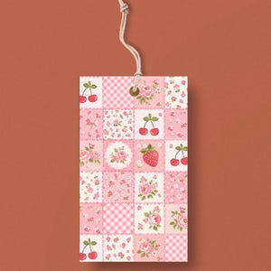 May include: A rectangular gift tag with a patchwork design featuring pink floral patterns, cherries, and a strawberry. The tag is made of paper and has a natural twine string attached for hanging.