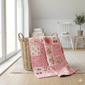 May include: A pink and white patchwork quilt with floral, strawberry, and cherry patterns draped over a wicker basket. The quilt features various square designs with roses, gingham, and fruit motifs. The basket has two handles.