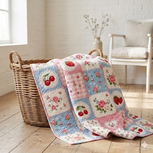 May include: A floral and fruit-patterned patchwork quilt with cherries, strawberries, and roses. The quilt is draped over a wicker basket. The quilt's colours are blue, pink, and white. The quilt is on a wooden floor.