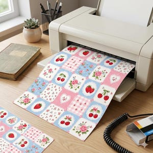 May include: A printer is shown printing a sheet of paper with a patchwork design. The design features squares with cherries, strawberries, roses, and gingham patterns in pink and blue. A second sheet of the same design is on the table.