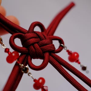 Könnte beinhalten: Nahaufnahme eines handgefertigten chinesischen Knotens in Burgunderrot. Der Knoten ist aufwendig mit einem dicken, strukturierten Kordel gewebt. Rote Perlen und kleine silberfarbene Charms hängen am Knoten und fügen ein dekoratives Element hinzu.