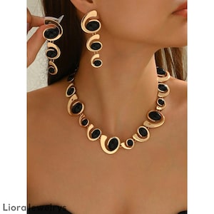 Puede incluir: Juego de joyas en tono dorado con collar y pendientes. El collar y los pendientes tienen un diseño repetitivo de piedras ovaladas negras engastadas en formas doradas y arremolinadas. Los pendientes son colgantes y el collar se coloca alrededor del cuello.