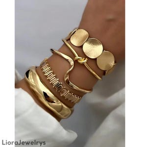Op de afbeelding: Een verzameling goudkleurige armbanden. De armbanden omvatten een dikke, afgeronde armband, een getextureerde band, een golvend ontwerp en een manchet met drie cirkelvormige accenten. De armbanden zijn op een pols gestapeld.