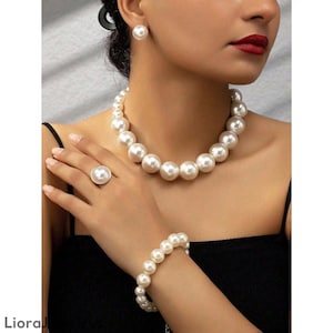 Può includere: Un set di gioielli in quattro pezzi con grandi perle finte bianche e rotonde. Il set comprende una collana, un braccialetto, un anello e orecchini a bottone. L'anello ha una fascia color oro. I gioielli sono indossati su un top nero.
