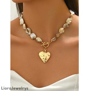 Puede incluir: Collar con colgante de corazón dorado y cierre de palanca. El colgante tiene una superficie texturizada y un diseño floral. El collar presenta cuentas irregulares, blanquecinas y marrones. La marca "LioraJewelrys" es visible en la parte inferior.