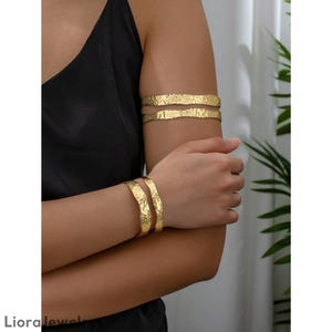Op de afbeelding: Goudkleurige arm- en polsmanchetten met een getextureerde, gehamerde uitstraling. De sieraden worden om de arm en pols gedragen. De manchetten hebben een ruwe, organische esthetiek en worden gepresenteerd tegen een zwarte top.