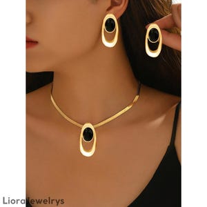Puede incluir: Juego de joyas doradas que incluye un collar, pendientes y un anillo. El conjunto incluye un colgante ovalado y pendientes con un centro de piedra negra. El anillo también tiene un diseño ovalado a juego.