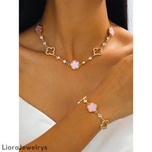Puede incluir: Un conjunto de joyas dorado con un collar y una pulsera. El collar y la pulsera tienen colgantes en forma de flor rosa y detalles de cristal transparente. El collar está alrededor del cuello y la pulsera en la muñeca.
