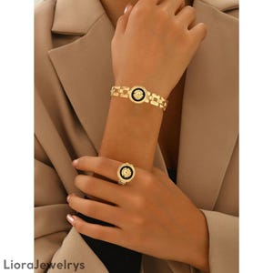 Puede incluir: Juego de pulsera y anillo dorados. La pulsera presenta una serie de eslabones rectangulares con un emblema circular negro y dorado. El anillo tiene un diseño similar. La joyería se usa en el brazo y la mano de una persona.