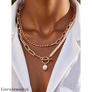 Può includere: Collana dorata a più strati con un design a catena, una catena di perline e una fila di perle. Include una chiusura a levetta con un pendente a perla pendente. Il marchio "LioraJewelrys" è visibile in basso.