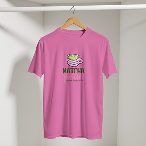 May include: A pink t-shirt with a matcha-themed design. The graphic shows a green matcha latte in a white cup, with the text "Matcha Girl" and "(Caffeine, but make it cute)". The shirt is displayed on a wooden hanger.