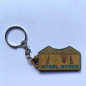 May include: Wooden keychain with a silver ring and chain. The keychain features a laser-cut design of a bridge with the words "STEEL RIVER" in blue. The keychain is made of light brown wood.