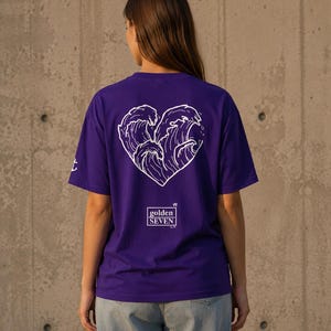 Könnte beinhalten: Ein violettes Kurzarm-T-Shirt mit einer weißen Linien-Grafik von Meereswellen in Herzform auf der Rückseite. Unter dem Herz befindet sich ein weißes Quadrat mit dem Text golden SEVEN. Ein kleines weißes Anker-Symbol ist auf dem Ärmel zu sehen.